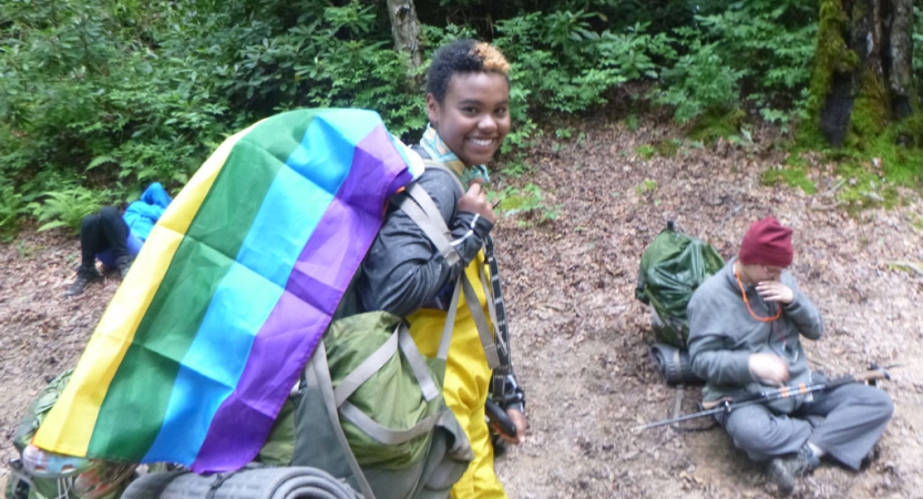 a backpacking student with a pride flag draped over their backpack smiles at the camera
